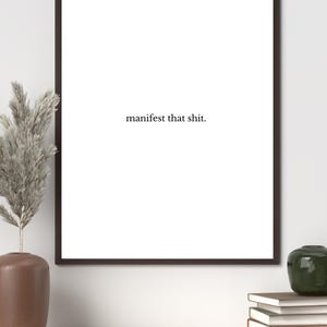 Könnte beinhalten: Ein gerahmter Druck mit dem Text "manifest that shit." Der Druck hat einen weißen Hintergrund und einen dunkelbraunen Rahmen. Eine braune Vase mit getrockneten Pflanzen, ein Stapel Bücher und ein grünes Dekorationsobjekt befinden sich auf einem Holzregal darunter.