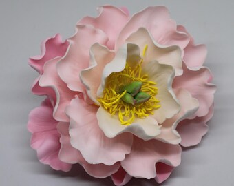 May include: A large, multi-layered artificial flower with soft pink, ruffled petals that transition to a lighter cream color toward the center. The center features bright yellow stamens surrounding two small, light green, bud-like structures.