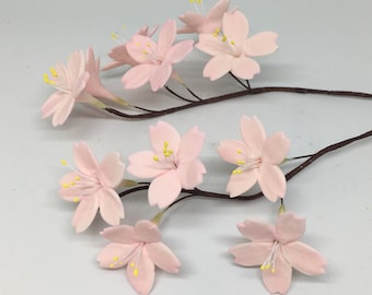 May include: A cluster of delicate pink cherry blossom flowers with yellow centers, arranged on brown stems. The flowers are made of fondant and are perfect for decorating cakes or cupcakes.