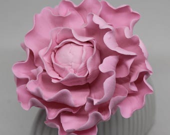 May include: A large, pink, artificial flower with multiple ruffled petals. The flower is a soft, pastel pink color and appears to be made of a pliable material. The flower is sitting on a white and gray striped vase.
