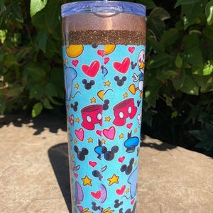 May include: A blue and white tumbler with a glitter top and a straw. The tumbler features a pattern of Mickey Mouse, Minnie Mouse, Donald Duck, and other Disney characters.
