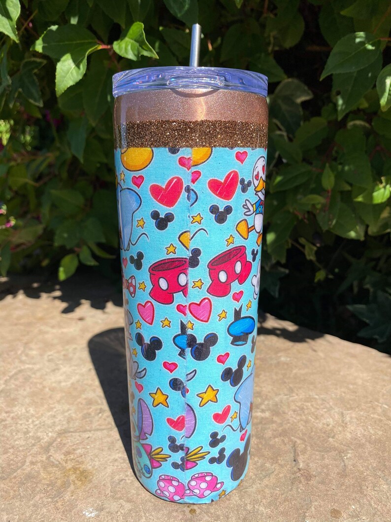 May include: A blue and white tumbler with a glitter top and a straw. The tumbler features a pattern of Mickey Mouse, Minnie Mouse, Donald Duck, and other Disney characters.