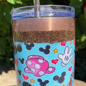 May include: A stainless steel tumbler with a brown glitter band and a blue and white pattern featuring Mickey Mouse and Minnie Mouse characters. The tumbler has a clear lid and a straw.