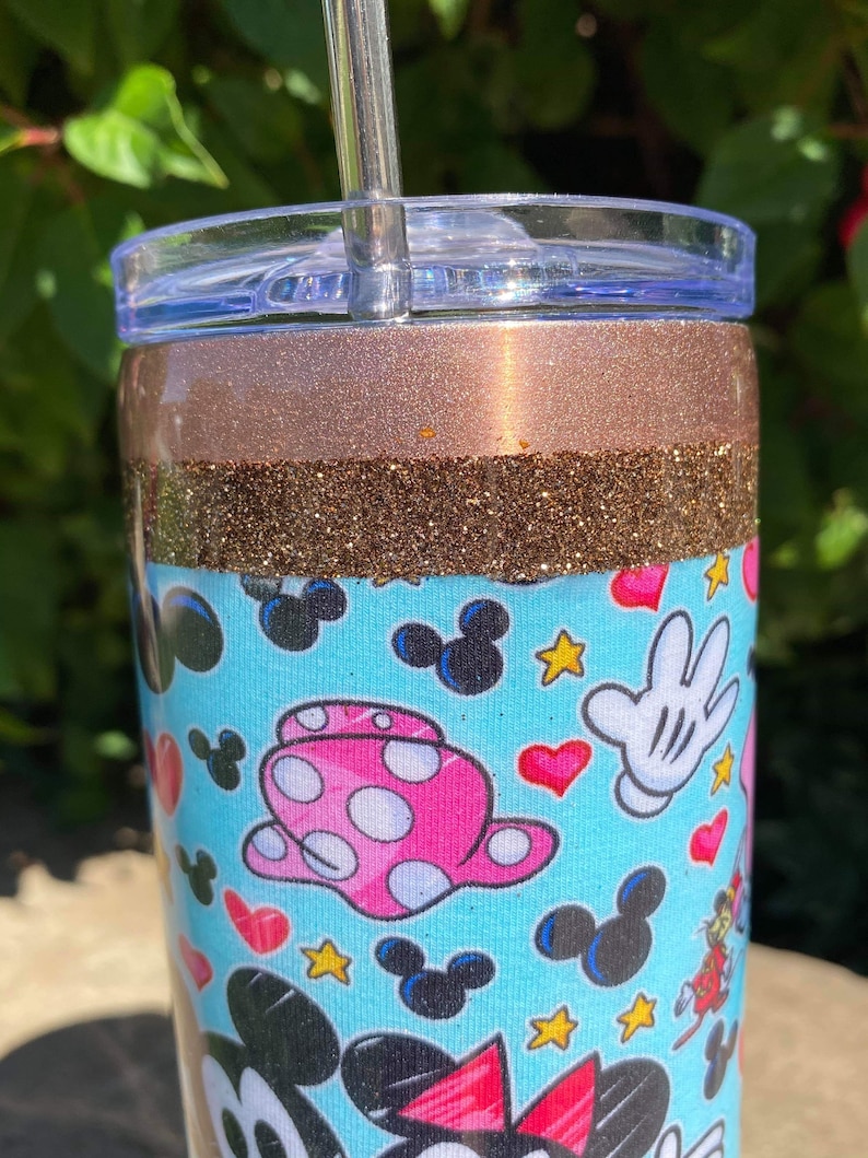 May include: A stainless steel tumbler with a brown glitter band and a blue and white pattern featuring Mickey Mouse and Minnie Mouse characters. The tumbler has a clear lid and a straw.