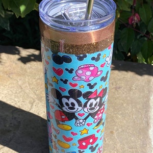 May include: A stainless steel tumbler with a blue and white design featuring Mickey Mouse and Minnie Mouse. The tumbler has a gold glitter band and a clear lid with a straw.