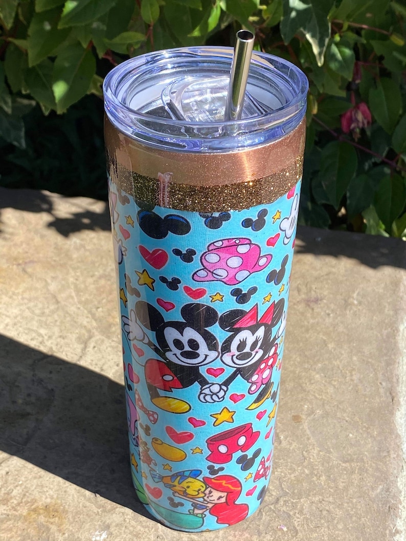 May include: A stainless steel tumbler with a blue and white design featuring Mickey Mouse and Minnie Mouse. The tumbler has a gold glitter band and a clear lid with a straw.