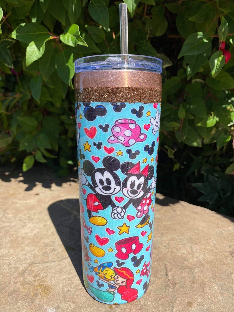 May include: A blue and brown tumbler with a straw. The tumbler has a glitter design and features a pattern of Mickey Mouse and Minnie Mouse characters, hearts, and stars.