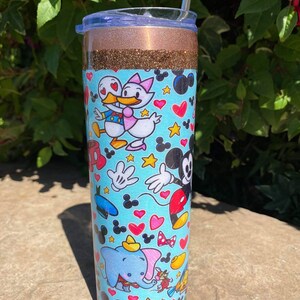 May include: A stainless steel tumbler with a gold glitter lid and a blue and white design featuring various Disney characters, including Mickey Mouse, Minnie Mouse, Donald Duck, and Dumbo. The tumbler has a straw.