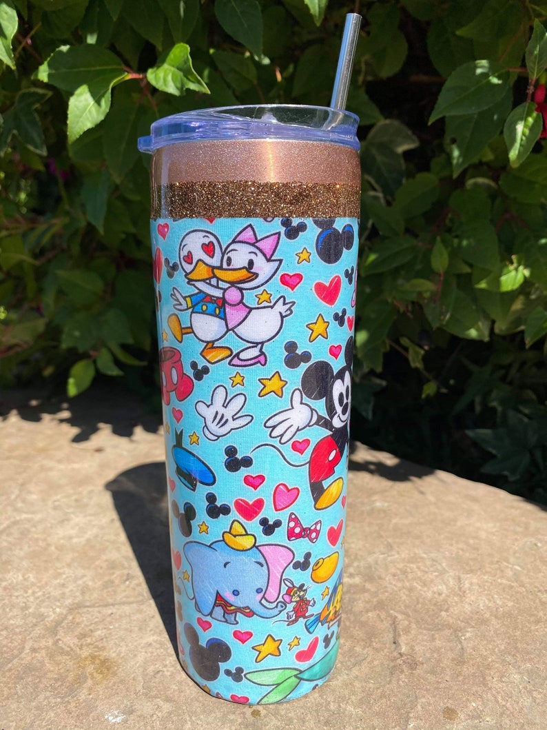 May include: A stainless steel tumbler with a gold glitter lid and a blue and white design featuring various Disney characters, including Mickey Mouse, Minnie Mouse, Donald Duck, and Dumbo. The tumbler has a straw.