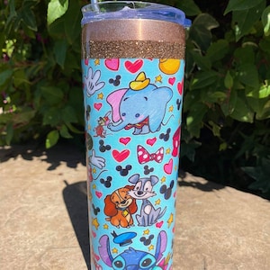 May include: A stainless steel tumbler with a blue and white design featuring various Disney characters, including Mickey Mouse, Stitch, and Lady and the Tramp. The tumbler has a gold glitter band and a clear lid.