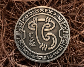 May include: A silver-colored metal coin with a circular design featuring a stylized symbol and text in an unknown language. The coin is surrounded by a decorative border.