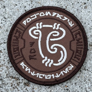 May include: A brown embroidered patch with a white symbol and text. The symbol is a stylized swirl with a line through the middle. The text around the symbol reads "TOOTONIV", "ESTAVIL", and "FUVNICTIV".