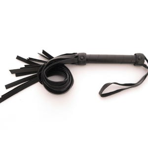 May include: A black leather whip featuring a cylindrical handle with a textured grip, a circular pommel, and a wrist strap. The whip includes a series of flat, flexible leather strips extending from the handle, with a coiled section of leather.
