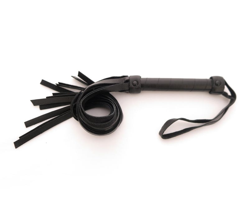 May include: A black leather whip featuring a cylindrical handle with a textured grip, a circular pommel, and a wrist strap. The whip includes a series of flat, flexible leather strips extending from the handle, with a coiled section of leather.