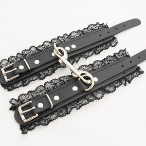 May include: A pair of black leather wrist cuffs with lace trim and silver metal hardware. The cuffs have a hook and loop closure.