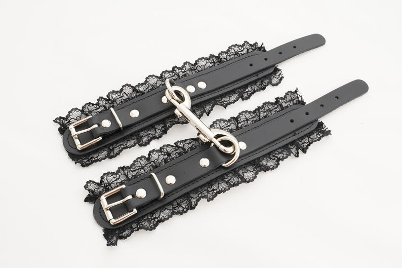 May include: A pair of black leather wrist cuffs with lace trim and silver metal hardware. The cuffs have a hook and loop closure.