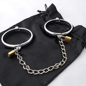 Op de afbeelding: Een paar zilveren metalen handboeien verbonden door een zilveren ketting met een messing slot. De handboeien liggen op een zwarte fluwelen tas.