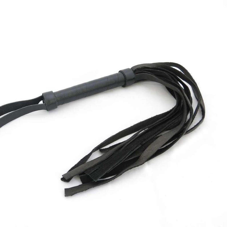 May include: A black leather whip featuring a cylindrical handle with a wrapped texture and a wrist loop at the end. The whip consists of multiple long, flat leather strips extending from the handle, creating a flexible and layered design.
