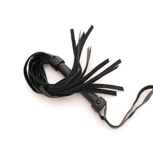 May include: A black leather flogger featuring a cylindrical handle with a wrist strap and multiple long, thin leather strips attached to the top. The strips are coiled around the handle, creating a layered, textured appearance in this item.