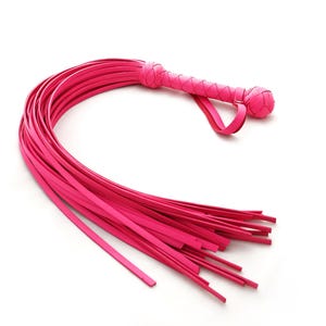 Puede incluir: Un látigo rosa brillante con un mango trenzado y un lazo. Las hebras del látigo son largas y finas, y se abren en abanico desde el mango. El mango tiene un extremo redondeado.