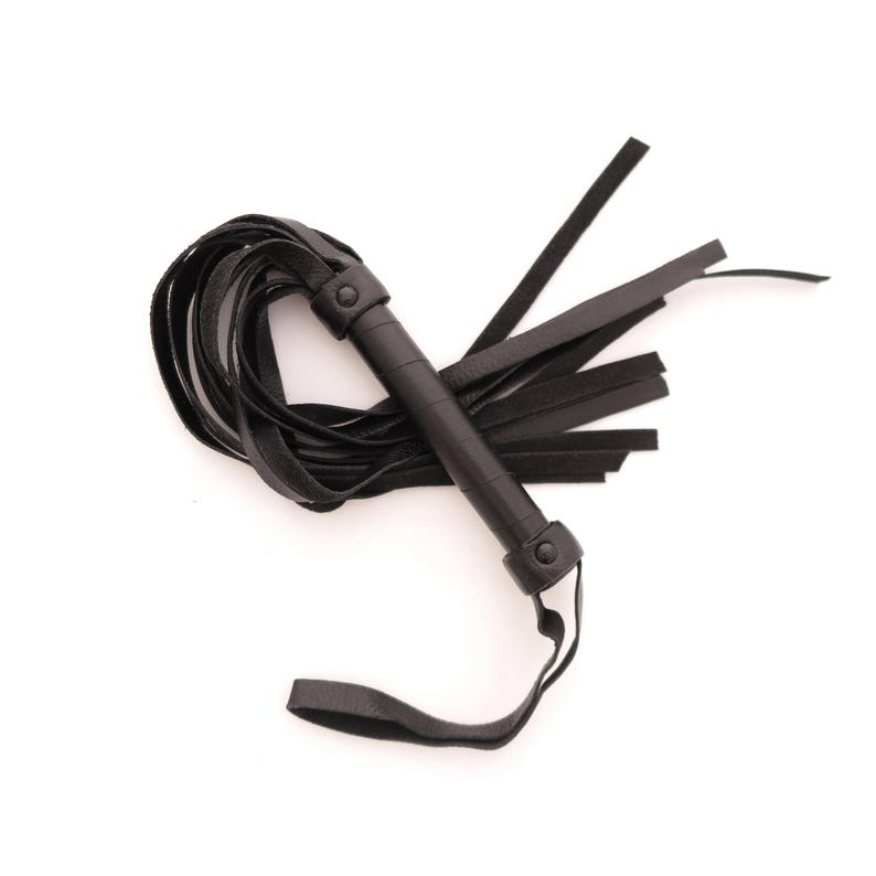 May include: A black leather flogger featuring a cylindrical handle wrapped in smooth leather, secured with two circular rivets at the top and bottom. Multiple long, flat leather strips extend from the handle, with a wrist loop attached at the base.