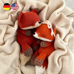 May include: Two handmade crochet fox plushies with orange bodies, white muzzles, and brown paws. One fox features a small cream-colored bow on its head. Both foxes have closed eyes and are positioned together, resting on a soft, textured cream fabric.