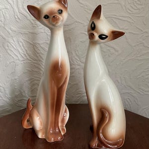 Op de afbeelding: Twee keramische kattenbeeldjes. De katten hebben een slank ontwerp met een witte basis en bruine accenten op de oren, het gezicht en de poten. Ze hebben zwarte ogen en zitten. De beeldjes staan op een donkerbruin oppervlak.