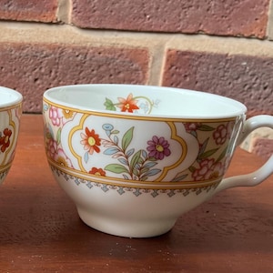 May include: A white teacup with a floral pattern of red, pink, and purple flowers, green leaves, and a yellow border. The teacup has a curved handle. The background is a brick wall.