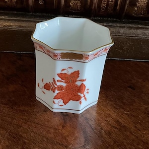 May include: White hexagonal porcelain vase with orange floral design and gold trim. The vase has a decorative band near the top and a brown base. The floral design is a stylized flower with leaves.