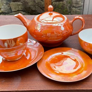 May include: An orange tea set with a pearlescent finish. The set includes a teapot, teacup, saucer, small bowl, and a small plate. The tea set is displayed on a wooden table.