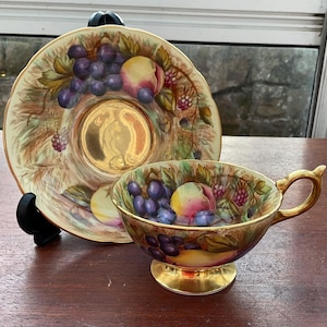Puede incluir: Un juego de taza y platillo de té vintage. La taza y el platillo están decorados con un diseño de frutas con uvas, melocotones y bayas. La taza tiene un asa y una base doradas. El platillo tiene un centro dorado.