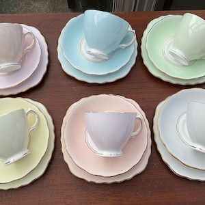 May include: Six pastel-colored teacups and saucers arranged on a brown surface. Each set includes a teacup and two saucers, with scalloped edges and gold trim. Colors include pink, blue, green, and yellow.