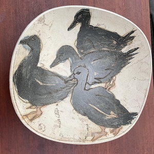 May include: A ceramic plate with a cream-colored background and a design of five stylized ducks in shades of gray and black. The ducks are arranged in a circular pattern, with their bodies overlapping. The plate has a slightly irregular shape.