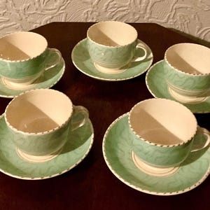 Op de afbeelding: Vijf bijpassende kopjes en schotels. De kopjes en schotels zijn lichtgroen met een witte binnenkant en een decoratief patroon. De randen zijn geschulpt met een gestippelde rand. De set staat op een donkerbruin oppervlak.