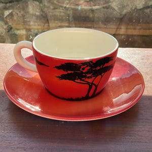Può includere: Un set di tazza e piattino in ceramica rossa. La tazza presenta una silhouette nera di un albero su sfondo rosso. Il piattino è rosso abbinato. La tazza ha un interno bianco e un manico curvo.
