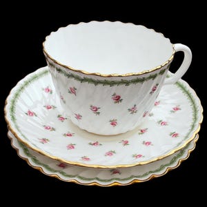 May include: A fine bone china teacup and saucer set. The white set features a pattern of small pink roses and a green border. The edges are trimmed with gold. The teacup has a handle and a fluted design. The saucer has a matching floral pattern.
