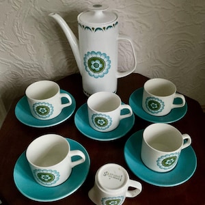 May include: A vintage white ceramic coffee set with a floral design in teal and green. The set includes a tall coffee pot, six cups, and saucers. The saucers and the floral design are teal. The cups have a white background.