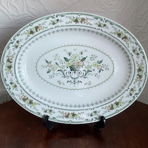 May include: Oval serving platter with a floral design. The white ceramic platter features a central oval with a basket of flowers and a scalloped border. The rim is decorated with a floral and leaf pattern in shades of green, yellow, and brown.