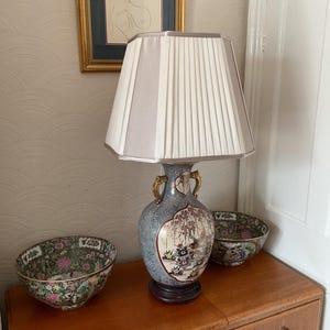 May include: A table lamp with a pleated lampshade in white and light grey stripes. The lamp sits on a wooden surface next to two decorative bowls with floral patterns. The lamp base is a blue and white vase with gold handles.