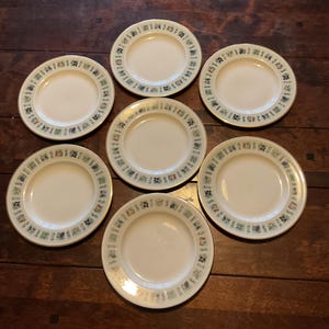 Op de afbeelding: Zeven witte keramische borden met een decoratieve rand van blauwe, groene en rode geometrische patronen. De borden zijn gerangschikt op een donker houten oppervlak. De borden zijn waarschijnlijk bedoeld om voedsel op te dienen.