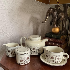 May include: A cream-colored tea set with a floral pattern. The set includes a teapot, creamer, sugar bowl, and a gravy boat with a saucer. The floral design features brown and white flowers. The set is displayed on a wooden surface.