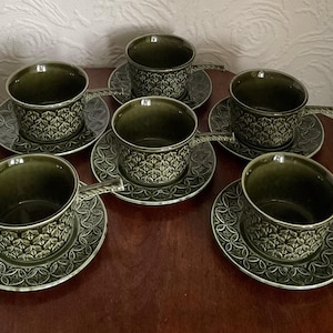 May include: Six olive green teacups and saucers. The cups have a textured pattern and a handle. The saucers have a matching design. The set is arranged on a dark wooden surface. A vintage tea set.