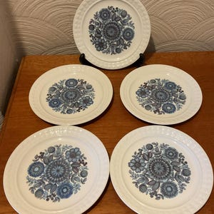 Può includere: Set di cinque piatti in ceramica bianca con un design floreale blu. I piatti hanno un bordo strutturato e un motivo floreale centrale nelle tonalità del blu e del grigio. Un piatto è esposto su un supporto nero.