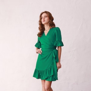 May include: A woman wearing a green wrap dress with ruffled sleeves and a ruffled hem. She is standing on a wooden floor with a red carpet in the background.