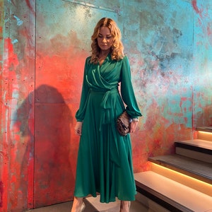May include: A woman wearing a long, emerald green dress with a wrap-style bodice and long sleeves. She is standing on a set of stairs with a textured wall behind her.