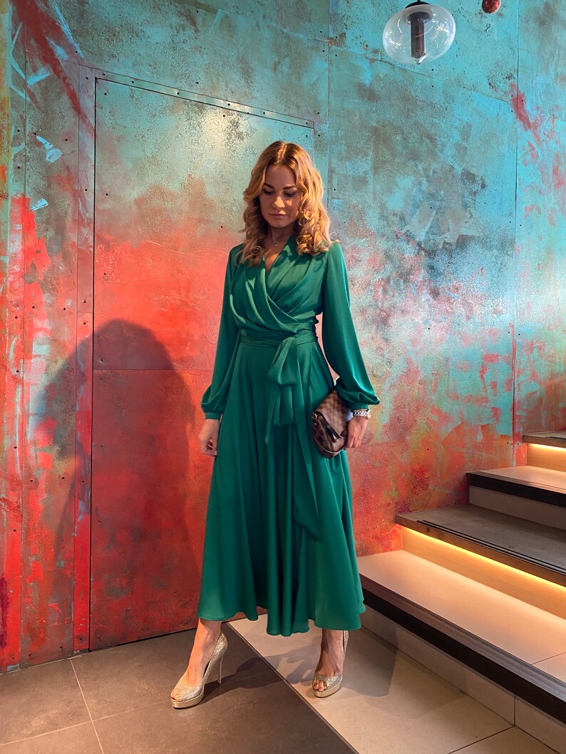 May include: A woman wearing a long, emerald green dress with a wrap-style bodice and long sleeves. She is standing on a set of stairs with a textured wall behind her.