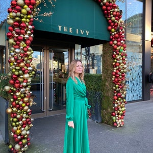 May include: A woman wearing a green satin dress and grey ankle boots stands in front of a building entrance decorated with a Christmas garland of red and gold baubles. The building has a green awning with the text "THE IVY" in gold lettering.