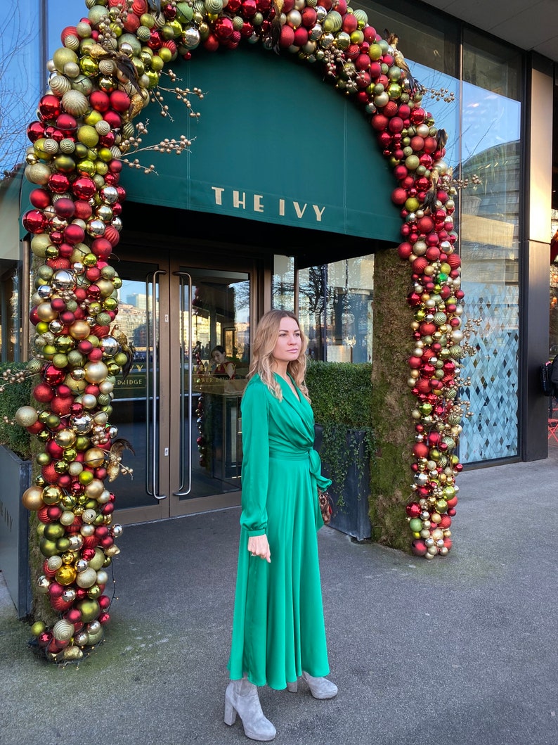 May include: A woman wearing a green satin dress and grey ankle boots stands in front of a building entrance decorated with a Christmas garland of red and gold baubles. The building has a green awning with the text "THE IVY" in gold lettering.