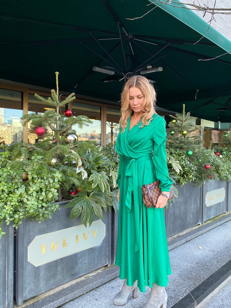 May include: A woman wearing a green wrap dress with a brown purse and grey boots. She is standing in front of a building with a sign that says "The Ivy".