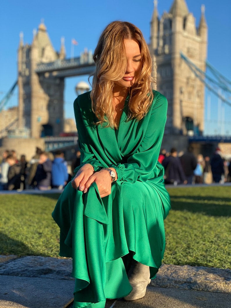 May include: A woman wearing a green satin dress sits in front of Tower Bridge in London. She is looking down at her hands, which are resting on her lap. The bridge is in the background, and the sky is blue.